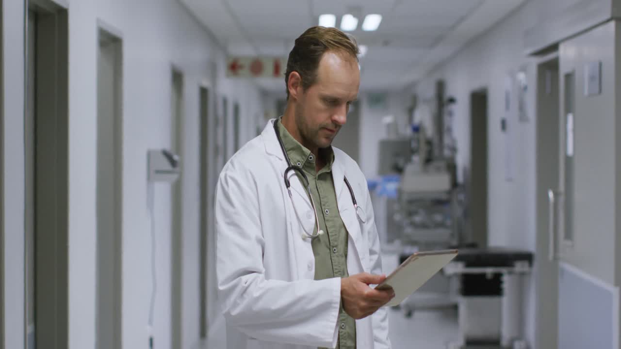 Ritratto di medico uomo caucasico che sorride mentre usa un tablet digitale nel corridoio dell'ospedale.