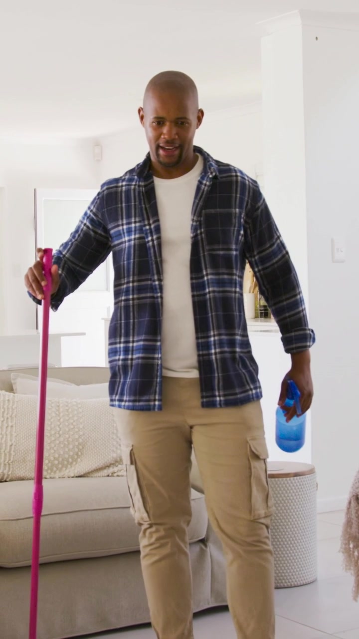Vertical video: Entering left, man in plaid holding pink mop and blue bottle, cleaning sofa at home