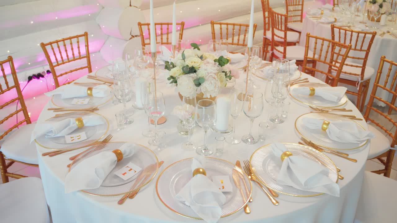 Et elegant dekket bryllupsbord med hvit duk, gullbestikk, blomsteroppsats og stearinlys, omgitt av gullstoler