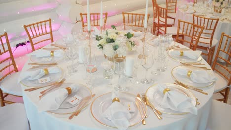 an elegant wedding table setup with white tablecloth, gold cutlery, floral centerpiece, and candles, surrounded by gold chairs.