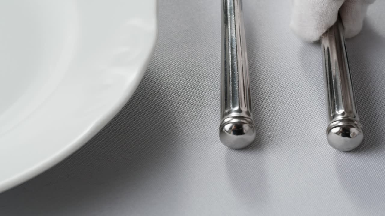 Professional table setting with white gloves. Plates, fork, knife, and glass arranged on a white table.