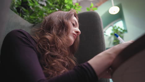 side view of young woman with long brown hair intensely focused while writing with blue pen, seated beside lush green plants in well lit modern setting, calm atmosphere and natural light present