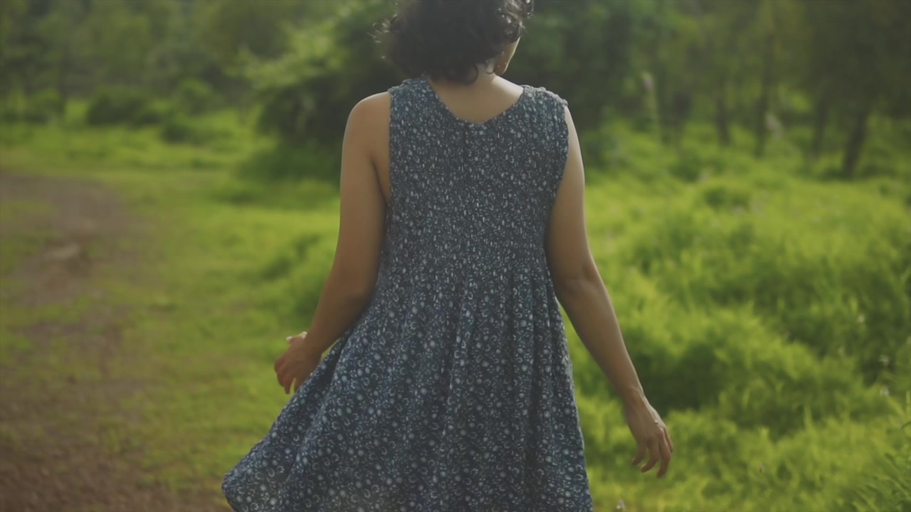 Female South Asian Model walking away from the camera wearing a blue flowing dress, towards a grassy forest