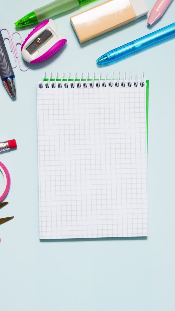 Portrait top down stop motion of colorful stationery circling a small grid notebook on light blue, leaving center copy space in a seamless loop