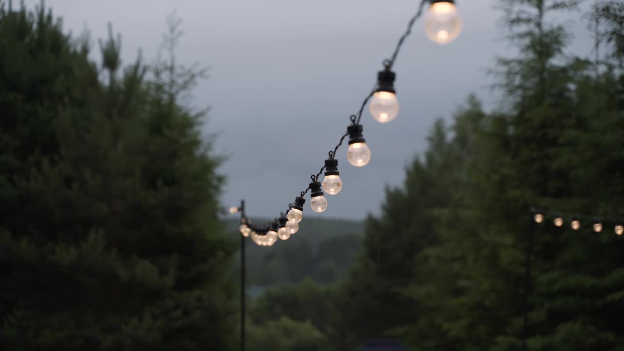 Garland of light bulbs hangs in yard. It is raining, water drops on light bulbs.