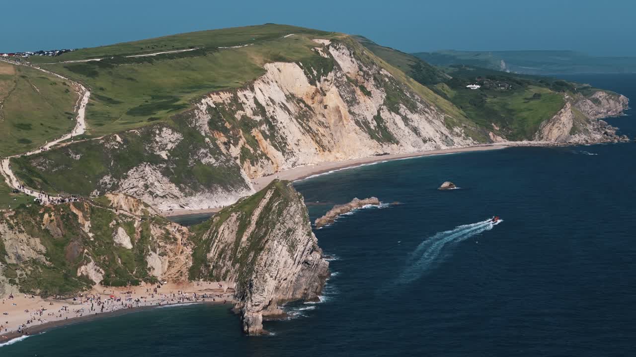 Drone tracks small boat along Dorset coastline with turquoise sea and rugged cliff edge below