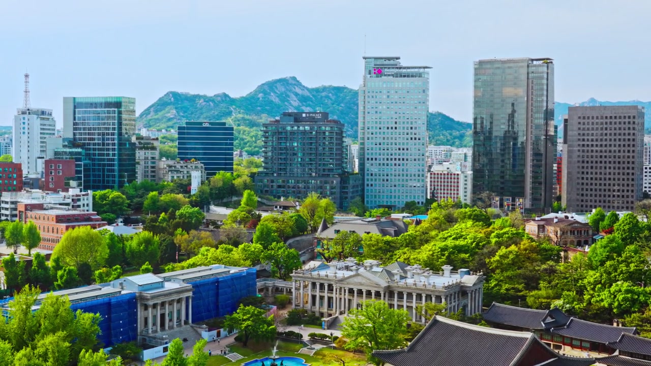 Istana Deoksugung menawarkan perpaduan unik arsitektur aula kayu tradisional Korea dan bangunan batu bergaya Barat, dikelilingi pepohonan rimbun dengan latar belakang cakrawala gedung pencakar langit modern Seoul.