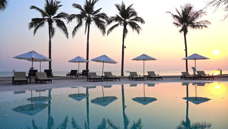 A-pink-and-yellow-tropical-sunset-behind-a-row-of-palm-trees,-empty-poolside-lounge-chairs,-and-beach-umbrellas