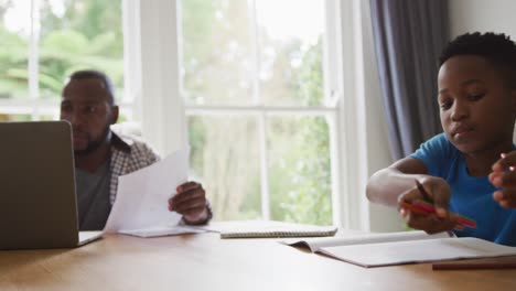 Afroamerikanischer-Mann,-Der-Zu-Hause-Mit-Einem-Laptop-Arbeitet-Und-Mit-Seinem-Sohn-Am-Tisch-Sitzt-Und-Schularbeiten-Erledigt