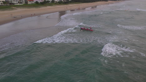 Luftaufnahme-Des-Mermaid-Beach-Slsc-Rennbootes,-Das-Während-Einer-Trainingseinheit-Mit-Der-Beliebten-Goldcoaststadt-Im-Hintergrund-Entlang-Meermaid-Beach-Goldcoast-Qld-Australien-An-Die-Küste-Kommt