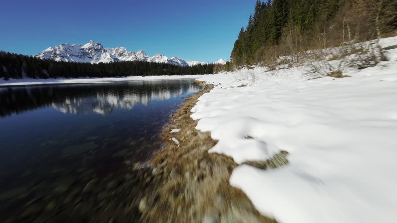 fotografía de un dron fpv del lado del lago cubierto de espesa nieve blanca con pinos y cordilleras en el fondo