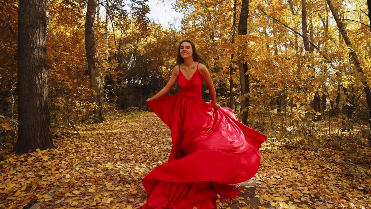 A graceful woman in a stunning flowing red dress walks through a vibrant autumn forest, surrounded by golden leaves, creating a picturesque scene of beauty and elegance