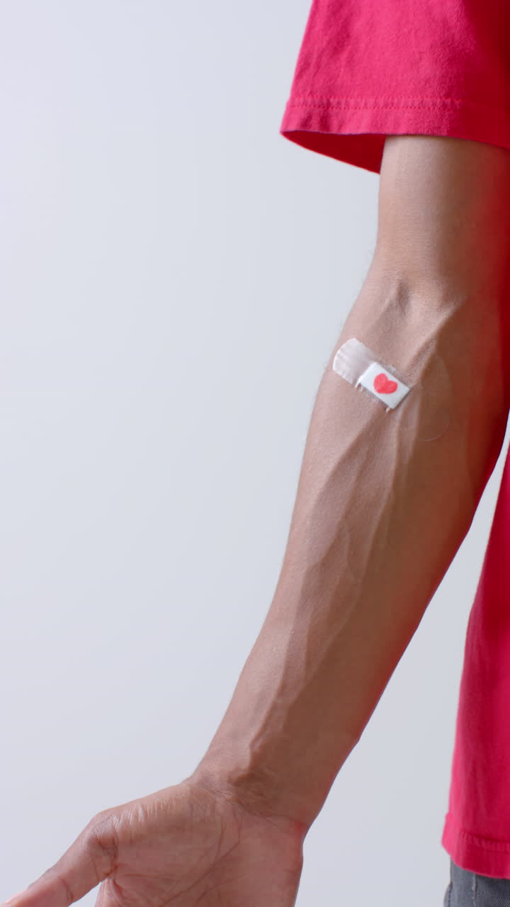 Vertical video of biracial man with red heart on bandage on his arm on white background, slow motion