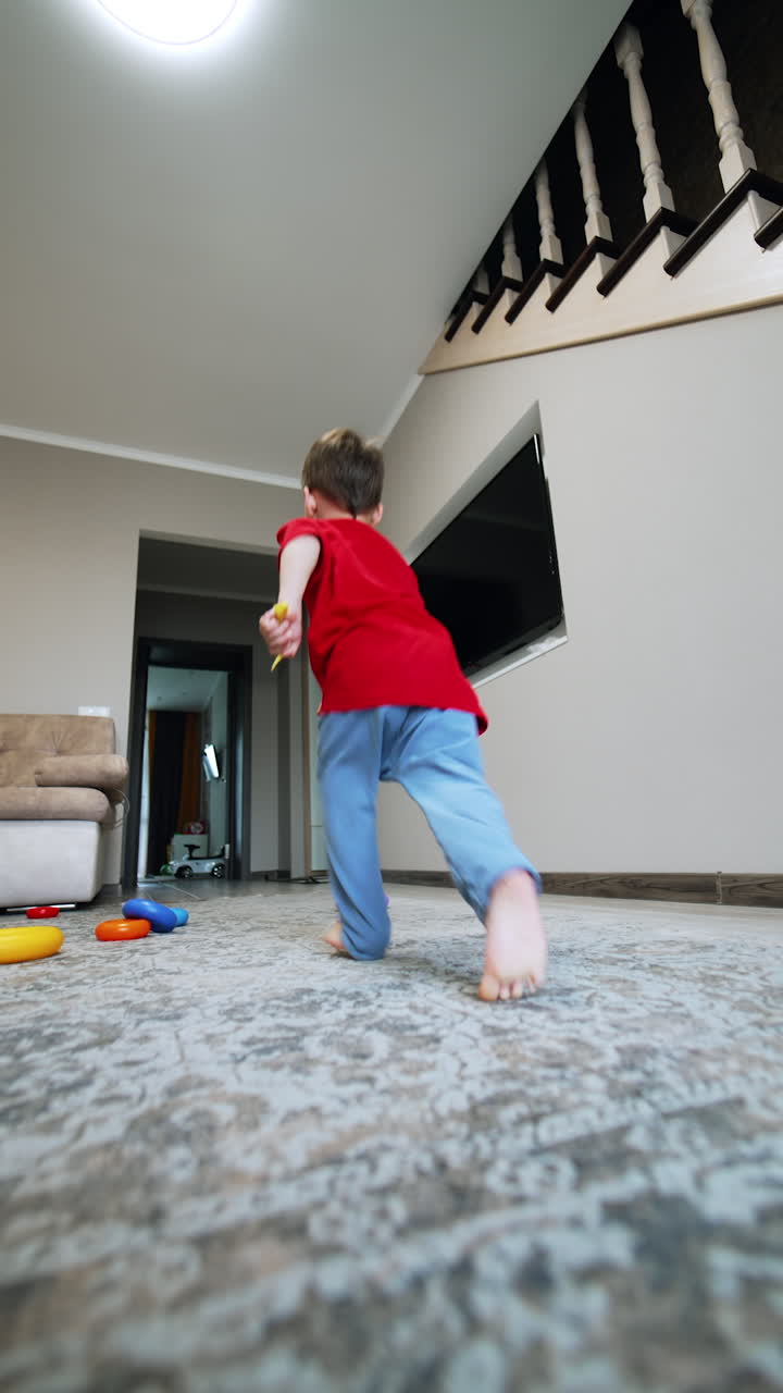 Caucasian baby boy in red t-shirt and blue pants enters the room. Kid throws a toy close to camera, comes to pick it up and runs away. Low angle view. Vertical video.