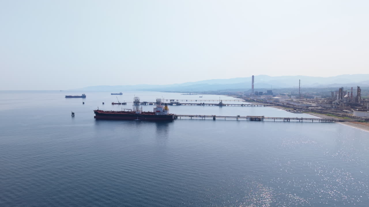 Drone ascends laterally from sea level showing oil tankers docked at refinery pier with distant mountains and industrial site on the Sicilian coast