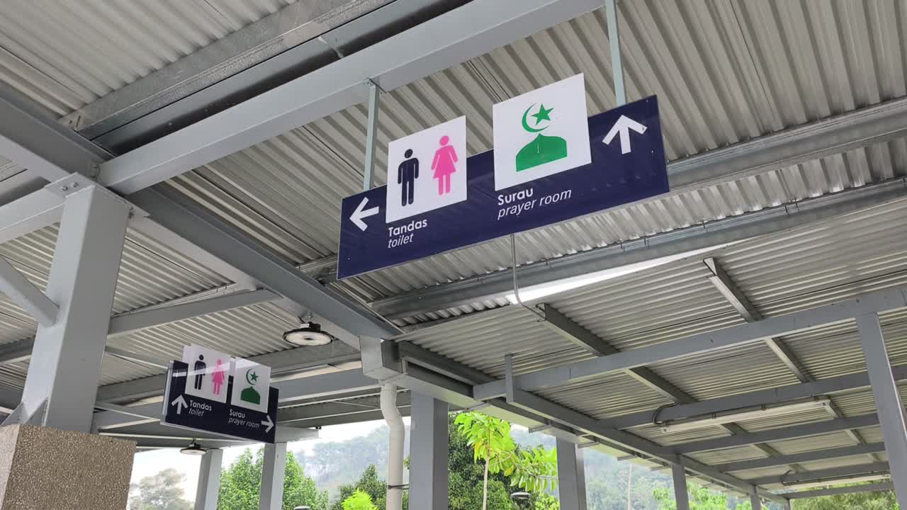 Papan petunjuk dwibahasa dalam bahasa Melayu dan Inggris mengarahkan pengguna ke toilet dan ruang salat. Ikon sederhana memastikan kejelasan. Papan tersebut digantung pada struktur tinggi untuk visibilitas maksimum