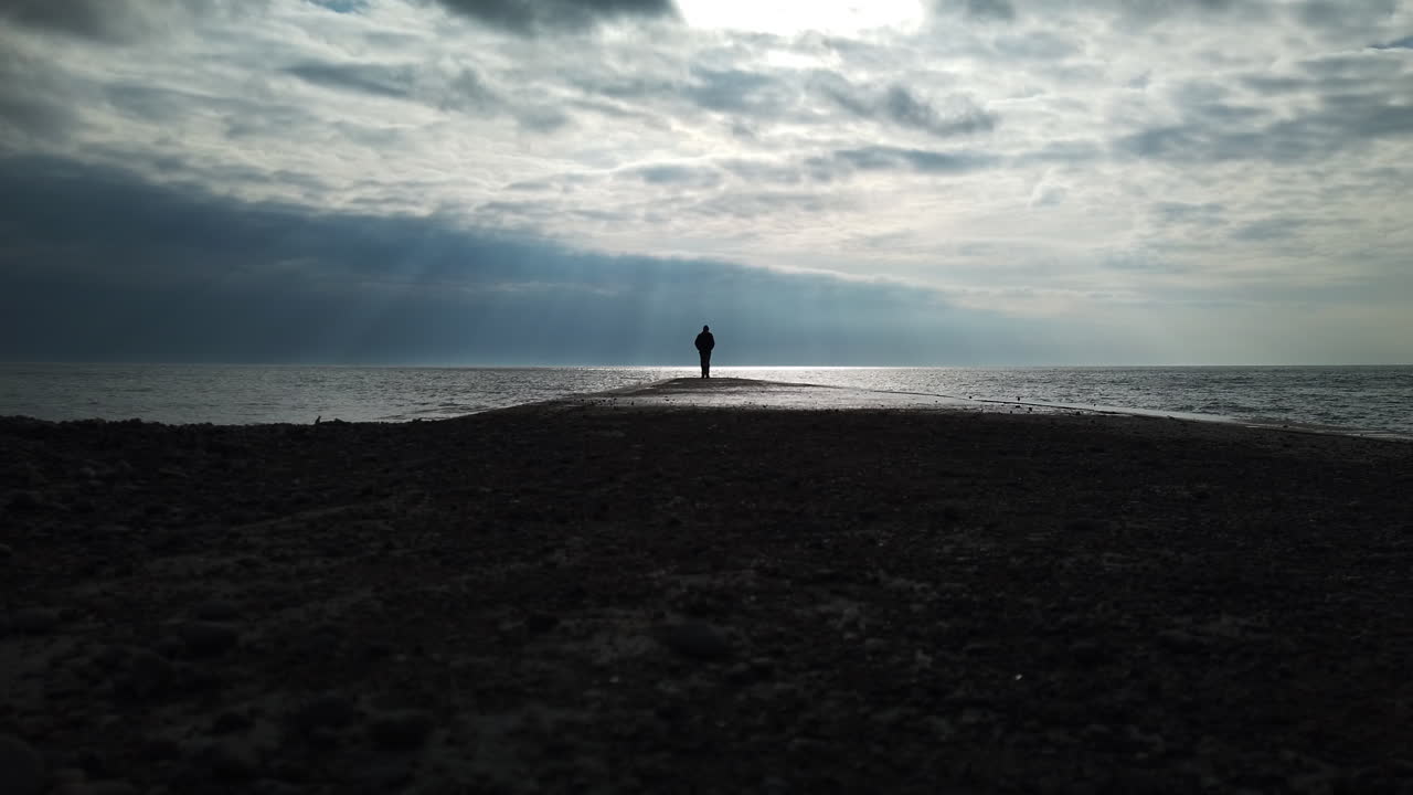 Man in a black coat leaving the jetty, walking towards camera, away from the horizon