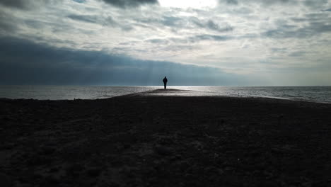 man in a black coat leaving the jetty, walking towards camera, away from the horizon