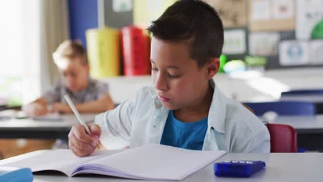 retrato de un niño de escuela caucásico feliz sentado en el aula, tomando notas, mirando a la cámara