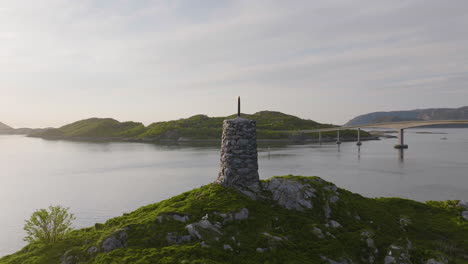 Rock-stacked-beacon-nearby-Sommaroy-Bridge,-sunset-aerial-arc,-Kvaloya