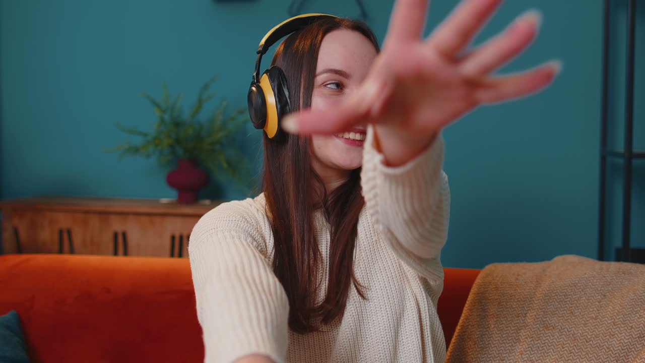 Giovane donna felicissima con cuffie wireless che balla e canta su un comodo divano nel soggiorno di casa.