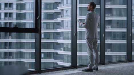 Thinking-man-executive-standing-using-tablet-working-online-in-window-office.