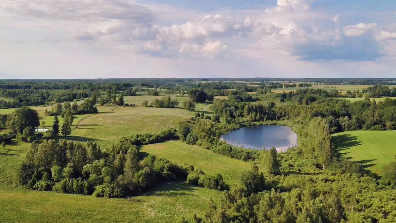 Lush green meadows and serene lakes create a picturesque landscape, captured from an aerial perspective under a cloudy sky, showcasing the natural beauty of the Latvian countryside