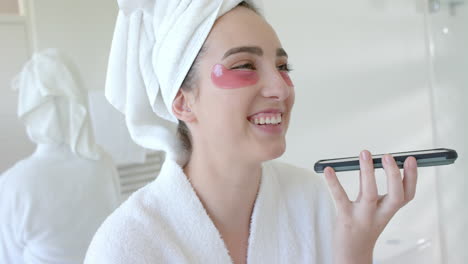 caucasian woman in bathrobe with towel on head and eye masks using phone in bathroom, slow motion