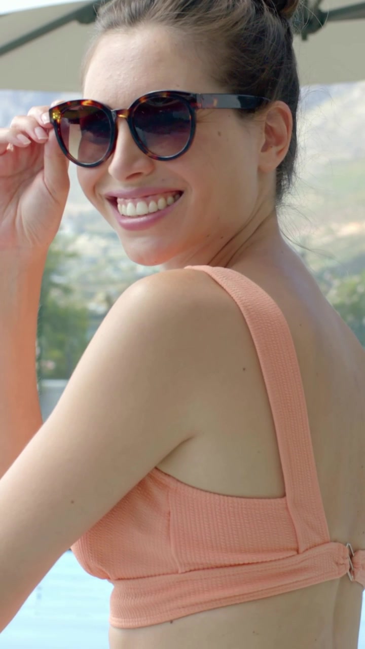 Video verticale: Il sole che causa abbagliamento, una donna che si gira e si sistema gli occhiali da sole a bordo piscina, con un top color pesca.