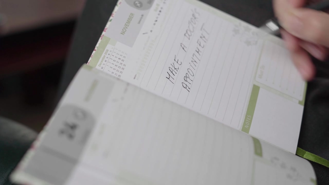 Female hand writes a doctor appointment reminder in a lined November day planner, noting personal health scheduling.