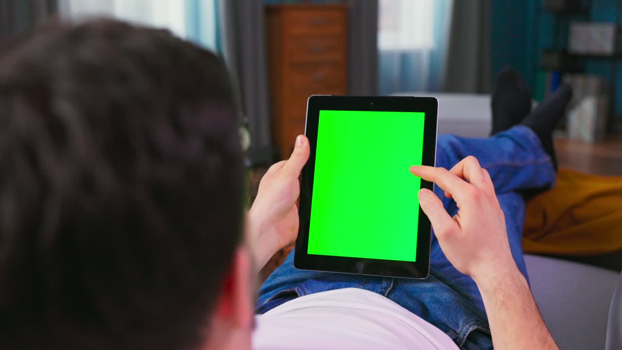 Young man at Home Resting on a Couch Using with Green Mock-up Screen Tablet Computer in in Vertical Portrait Mode. Boy Using Gestures with Touchscreen Device, Browsing Internet, Watching Content