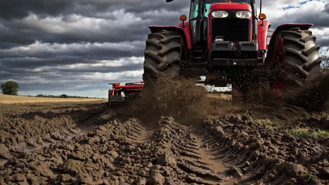 red tractor plowing a muddy field under a cloudy sky