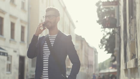 stylish man standing in the old town street in sunlight and talking on the mobile phone, then looking at the watch and going away