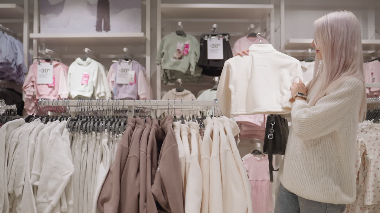 Elegant lady browsing soft polo shirts on rack in store gently feeling fabric texture as she inspects garment quality with delicate hand movements amidst pastel clothing display and retail ambiance