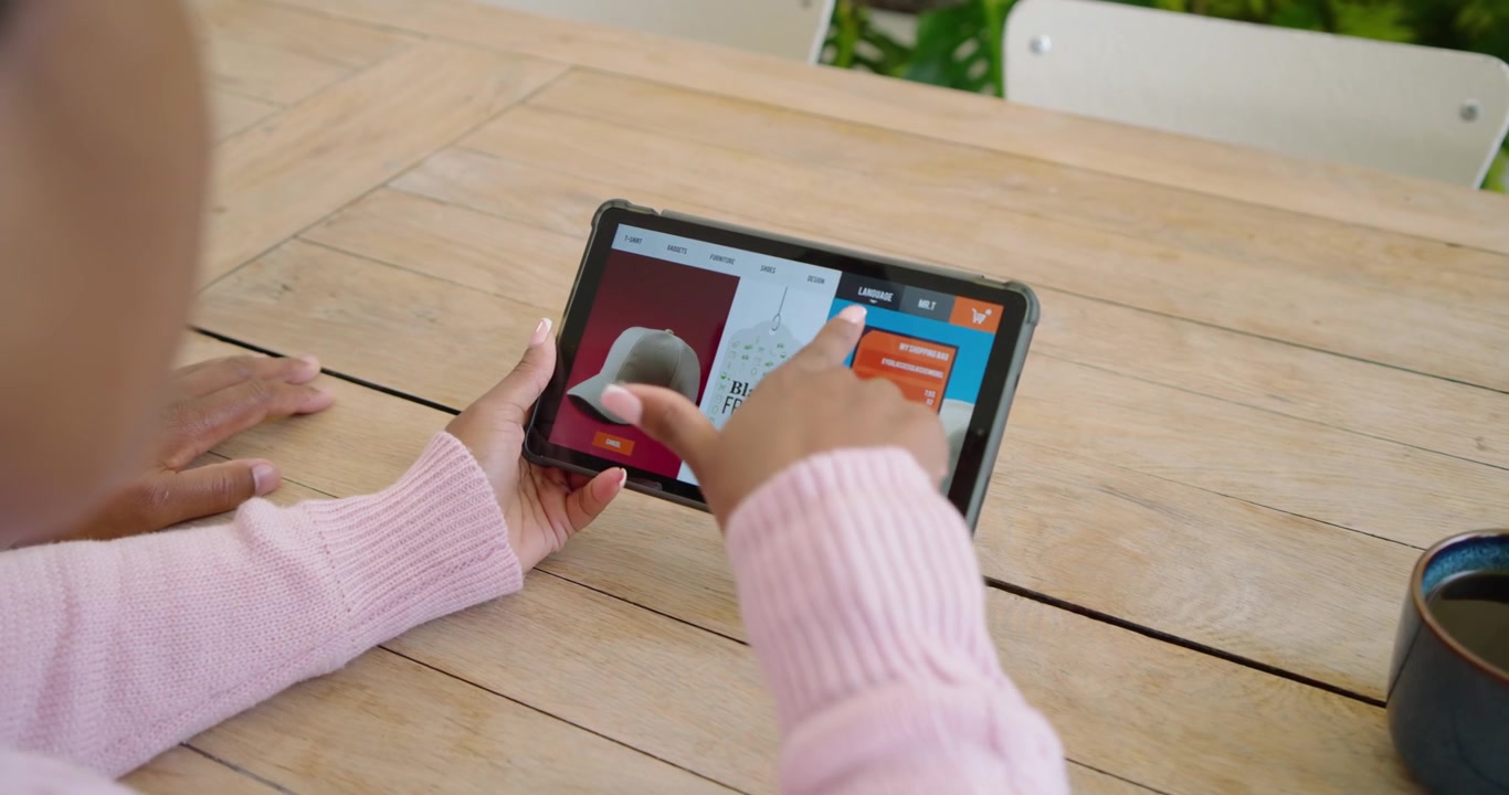 African woman seeing Black Friday hat deal leaning at wooden table swiping tablet as man pointing