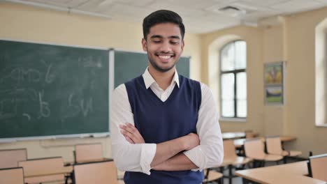 portrait of a young male teacher smiling with crossed arms, standing in a classroom in front of a blackboard, showing confidence and passion for education