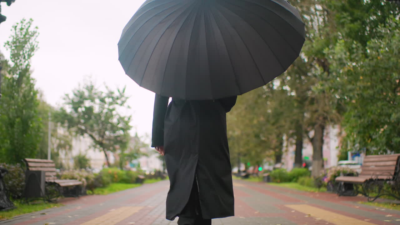 Person in long coat walking under large black umbrella along tree-lined park pathway on overcast day, benches and vintage lampposts on both sides, evoking calm mood of autumn solitude and urban lifestyle