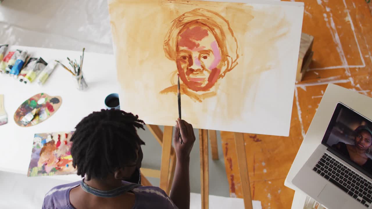 Artista afroamericano con mascherina che dipinge su tela e guarda il laptop nello studio d'artista.