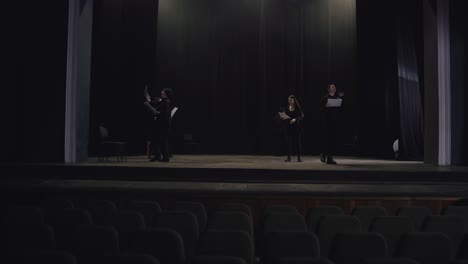 view from afar a confident group of young actors in black suits walk around the stage with sheets of paper in their minds reading their text and script of the play and preparing for their performance on the stage of a theater with a black curtain