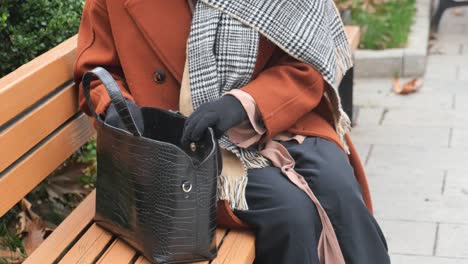 woman in a red coat and black gloves sitting on a bench looking into her black bag