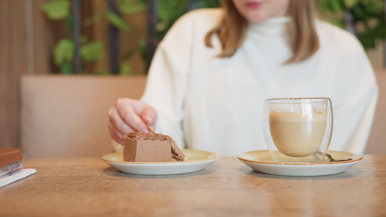 close up of woman in white sweater gently cutting chocolate cake on ceramic plate with fork, latte in double walled glass beside, soft cafe lighting