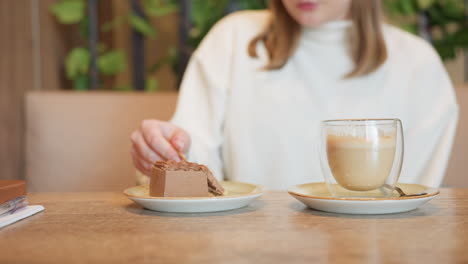 close up of woman in white sweater gently cutting chocolate cake on ceramic plate with fork, latte in double walled glass beside, soft cafe lighting