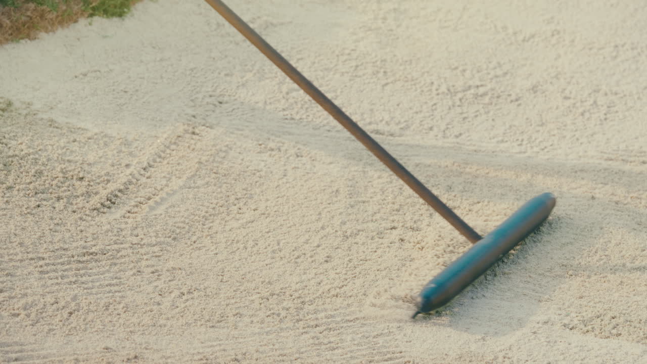 Slow motion closeup of a rake smoothing and grooming sand in a golf bunker. No people visible. Bright sunny day on a well-maintained golf course, capturing the texture and care of course maintenance.