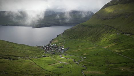 the serene village of funnings in the faroe islands rests in a green valley, framed by winding roads, steep hills, and a peaceful ocean inlet under dramatic, cloud-filled skies