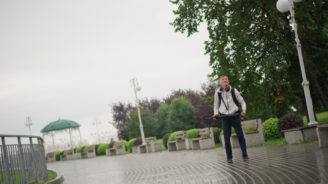 바닷가 산책로에서 바람 때문에 우산과 씨름하며, 강풍에 맞서 우산을 접고 백팩을 고정하고, 마치 통근자에서 여행자이자 거리 공연가가 된 듯한 모습으로 흠뻑 젖은 재킷을 입고 있는 백인 남성