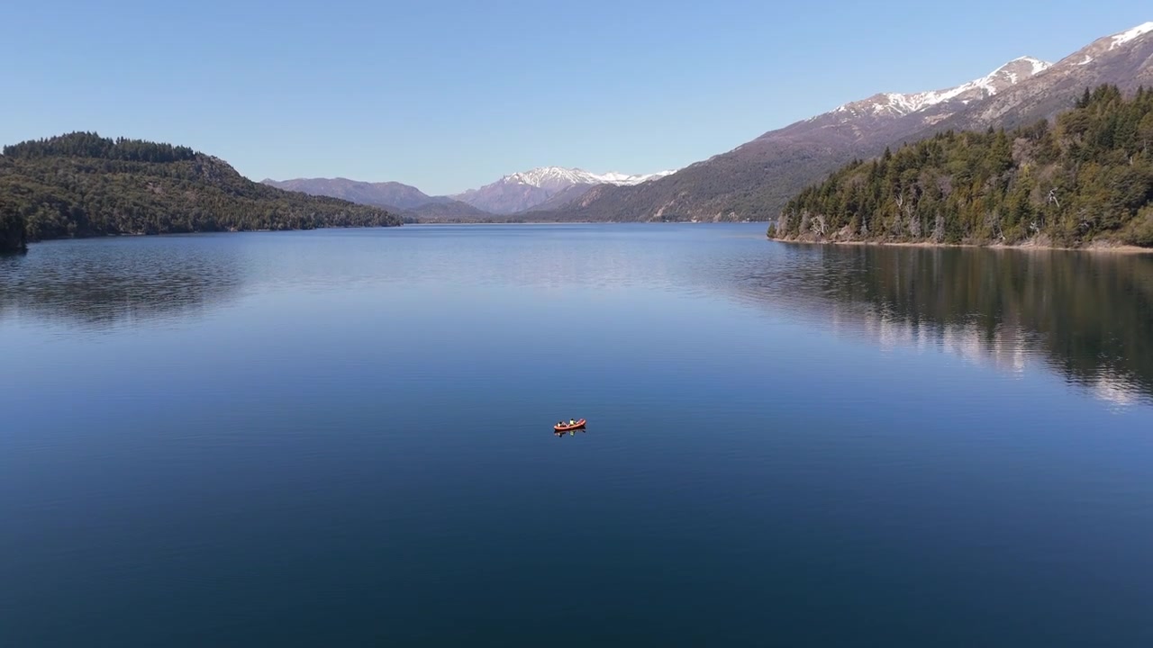 Vista aérea em movimento de pan para baixo de um lago com um esportista ou homem remando de caiaque sozinho, com paisagem florestal e montanhas nevadas em Bariloche, Rio Negro, Argentina.