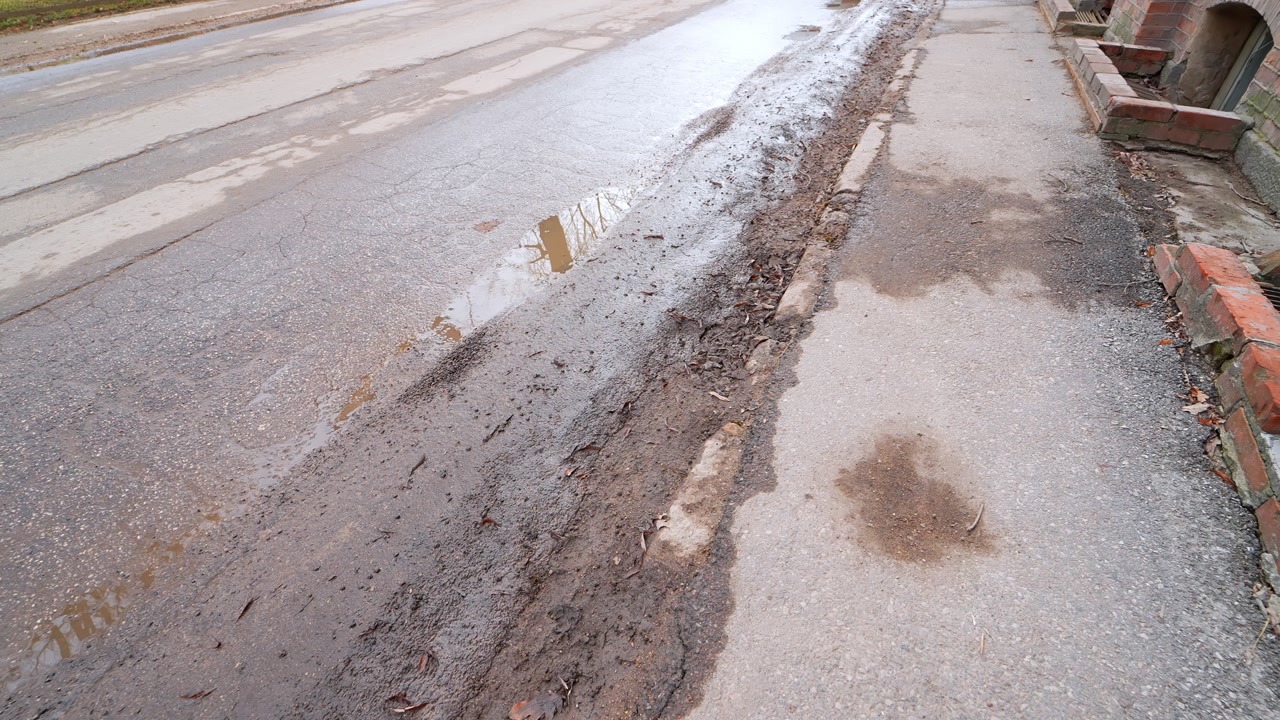 Curbside slush and muddy runoff beside a wet street as a car passes—spring thaw mess with puddles, grit, and cracked pavement after winter road salt.