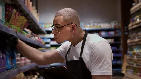 Arbeiter-Gemischter-Abstammung-In-Schürze,-Der-Im-Supermarkt-Auf-Dem-Regal-Waren-Mit-Tablet-Zählt