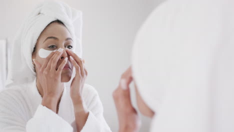 Biracial-woman-applying-under-eye-patches-looking-in-mirror-in-bathroom,-slow-motion