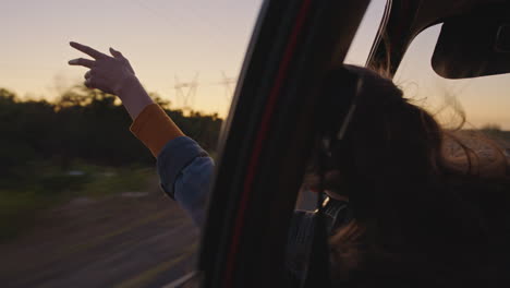 Frau-Im-Auto-Hält-Die-Hand-Aus-Dem-Fenster-Und-Spürt,-Wie-Der-Wind-Durch-Die-Finger-Weht,-Während-Sie-Auf-Einem-Roadtrip-Durch-Die-Landschaft-Fährt-Und-In-Den-Sommerurlaub-Reist-Und-Bei-Sonnenaufgang-Die-Freiheit-Auf-Der-Straße-Genießt